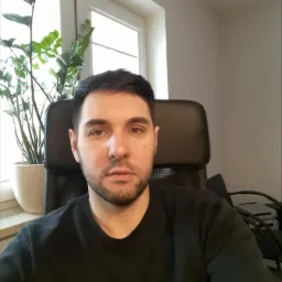 Portret mężczyzny siedzącego na krześle biurowym, w tle okno z rośliną doniczkową, neutralne oświetlenie.