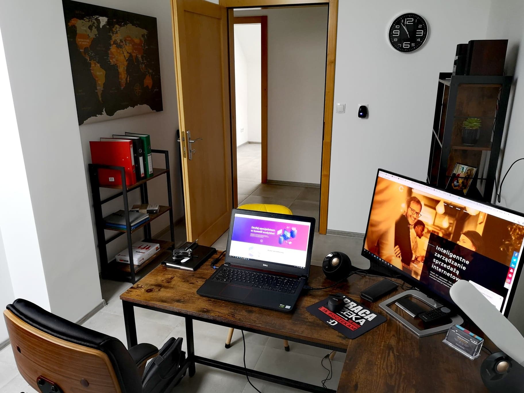 Stanowisko pracy z dwoma monitorami wyświetlającymi strony internetowe, laptop Dell, podkładka pod mysz z napisem 'Praca czeka, ja poczekam', zegar na ścianie, biurko z blatem imitującym drewno...