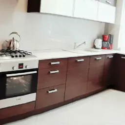 Nowoczesna kuchnia z ciemnobrązowymi, drewnopodobnymi frontami szafek, białym blatem i białymi górnymi szafkami, wyposażona w srebrny czajnik, płytę gazową i piekarnik, z logo firmy w prawym dolnym...