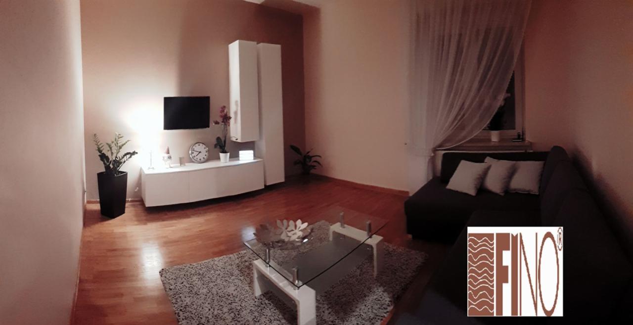 Nowoczesny salon z białymi szafkami RTV, szarą sofą narożną, szklanym stolikiem kawowym na puszystym dywanie i drewnianą podłogą, oświetlony ciepłym światłem.