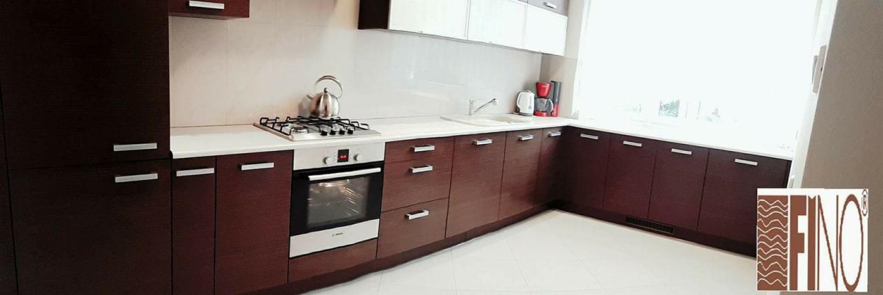 Nowoczesna kuchnia z ciemnobrązowymi, drewnopodobnymi frontami szafek, białym blatem i białymi górnymi szafkami, wyposażona w srebrny czajnik, płytę gazową i piekarnik, z logo firmy w prawym dolnym...