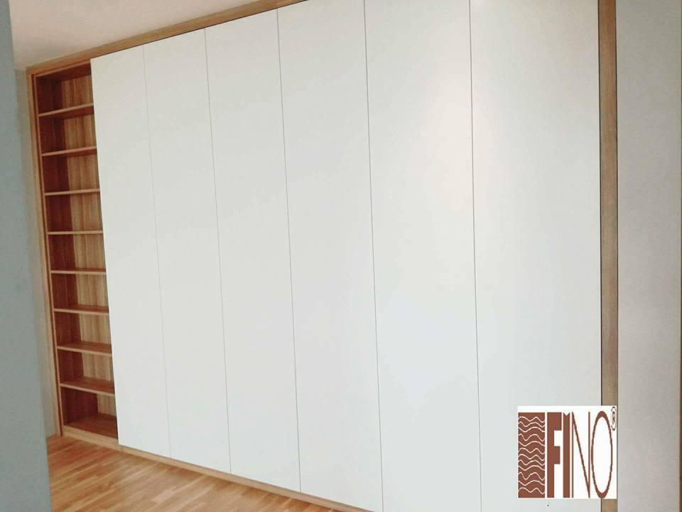 Duża, biała szafa z przesuwnymi drzwiami i drewnianymi półkami po lewej stronie, wbudowana w ramę z jasnego drewna.