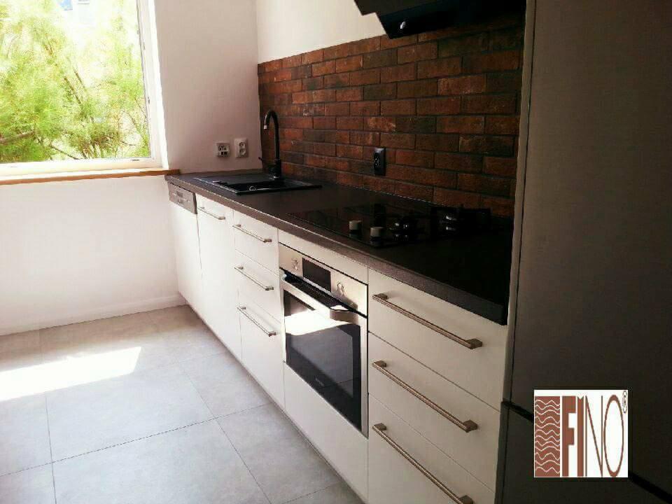 Nowoczesna kuchnia z białymi frontami, czarnym blatem i ścianą z cegły, widok na okno z zielenią na zewnątrz.