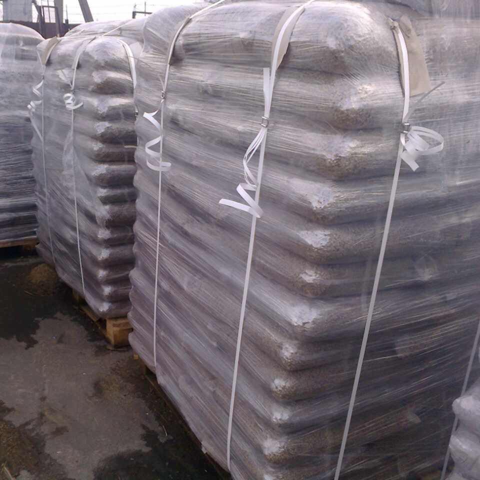 Sztaple worków z granulatem, owinięte folią i zabezpieczone białymi paskami, ułożone na paletach na zewnątrz.