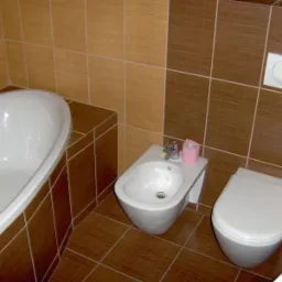 Ukończona łazienka z brązowo-beżowymi płytkami, zawierająca wannę z obudową z płytek, bidet z rolką różowego papieru toaletowego i podwieszaną toaletę z przyciskiem spłukiwania.