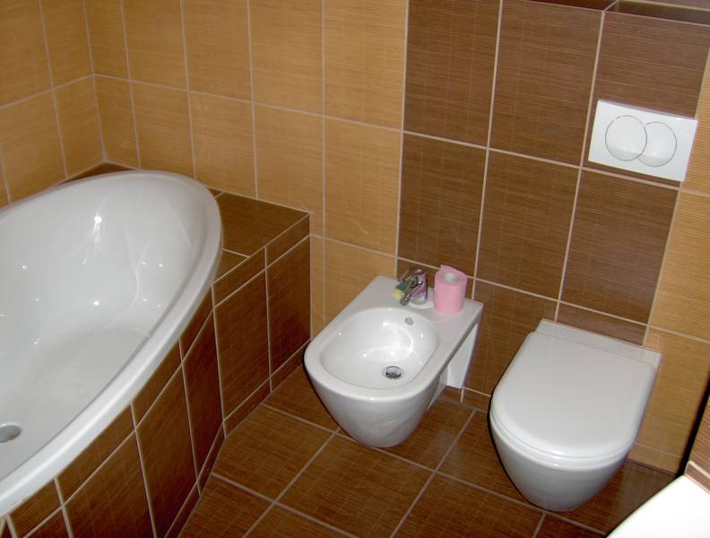 Ukończona łazienka z brązowo-beżowymi płytkami, zawierająca wannę z obudową z płytek, bidet z rolką różowego papieru toaletowego i podwieszaną toaletę z przyciskiem spłukiwania.