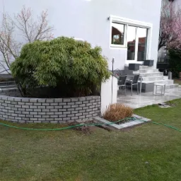 Kamienny taras z jasnoszarych płyt prowadzący do domu, z niskim, półokrągłym murem z cegieł i bambusem w centralnej części ogrodu.