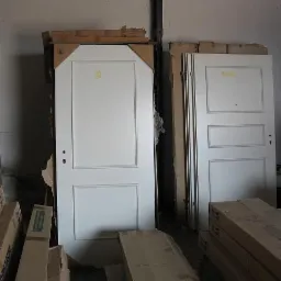 Białe, dwuskrzydłowe drzwi wewnętrzne z panelami, stojące w magazynie obok kartonów z framugami i innymi drzwiami.