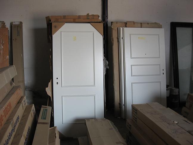 Białe, dwuskrzydłowe drzwi wewnętrzne z panelami, stojące w magazynie obok kartonów z framugami i innymi drzwiami.