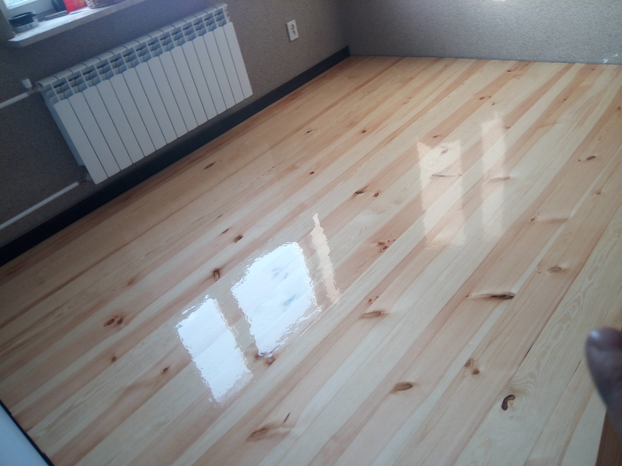 Błyszcząca, jasna drewniana podłoga z widocznym grzejnikiem na ścianie, po renowacji.