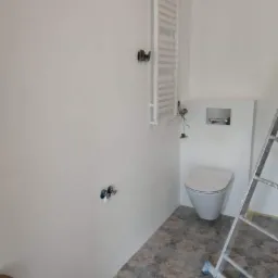 Biała łazienka w trakcie wykańczania: zamontowany grzejnik drabinkowy, częściowo podłączona toaleta, widoczne wystające kable, przygotowane przyłącza wodne, wzorzysta podłoga i drabina.