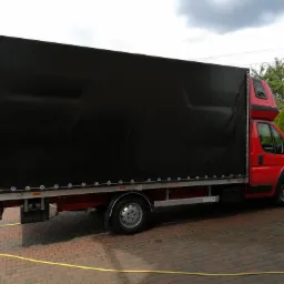 Czerwony samochód dostawczy z czarną plandeką na tle brukowanej nawierzchni i zieleni.