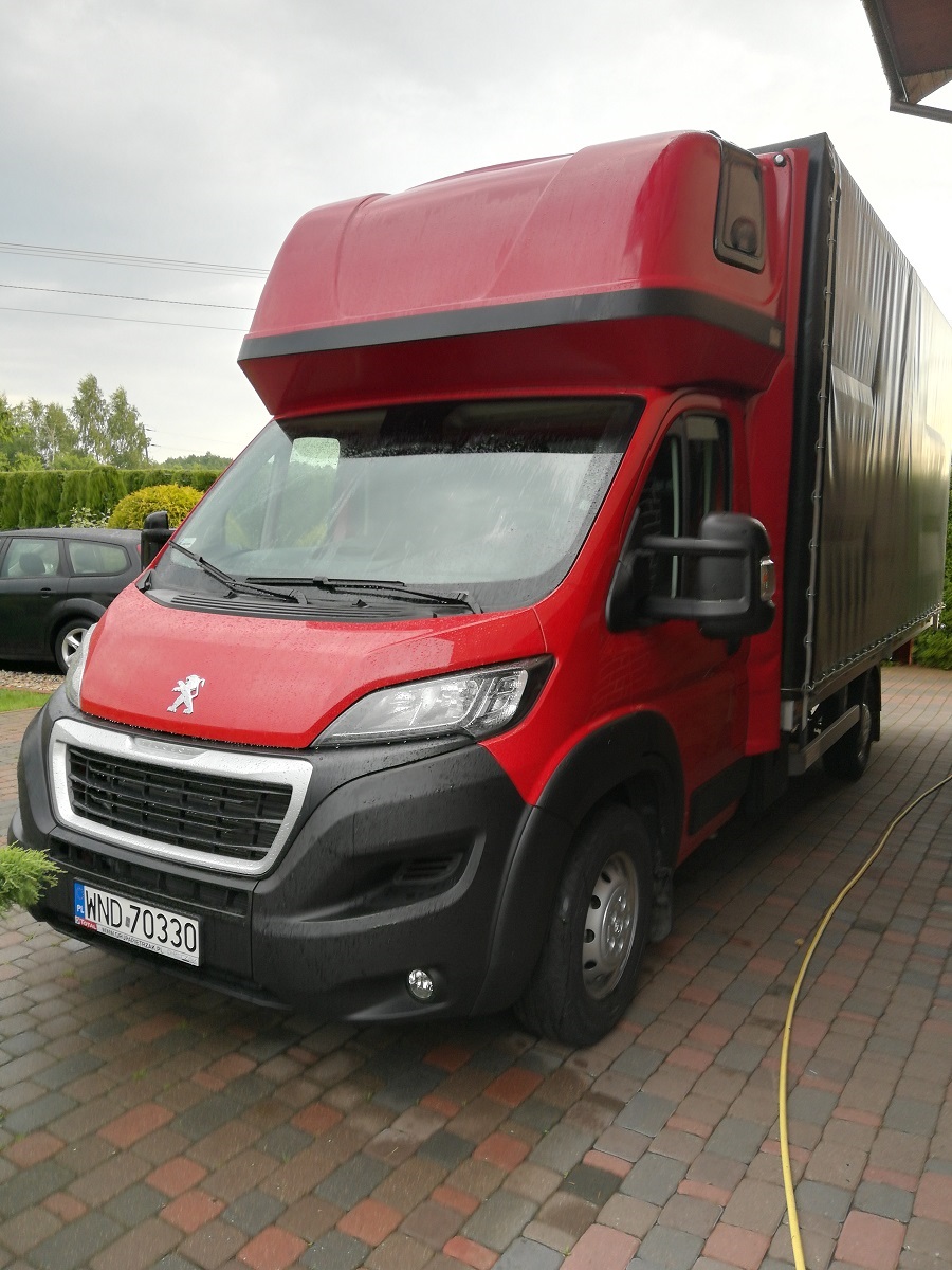 Czerwony samochód dostawczy Peugeot z plandeką, zaparkowany na brukowanym podjeździe w deszczowy dzień, widoczne tablice rejestracyjne WND 70330.