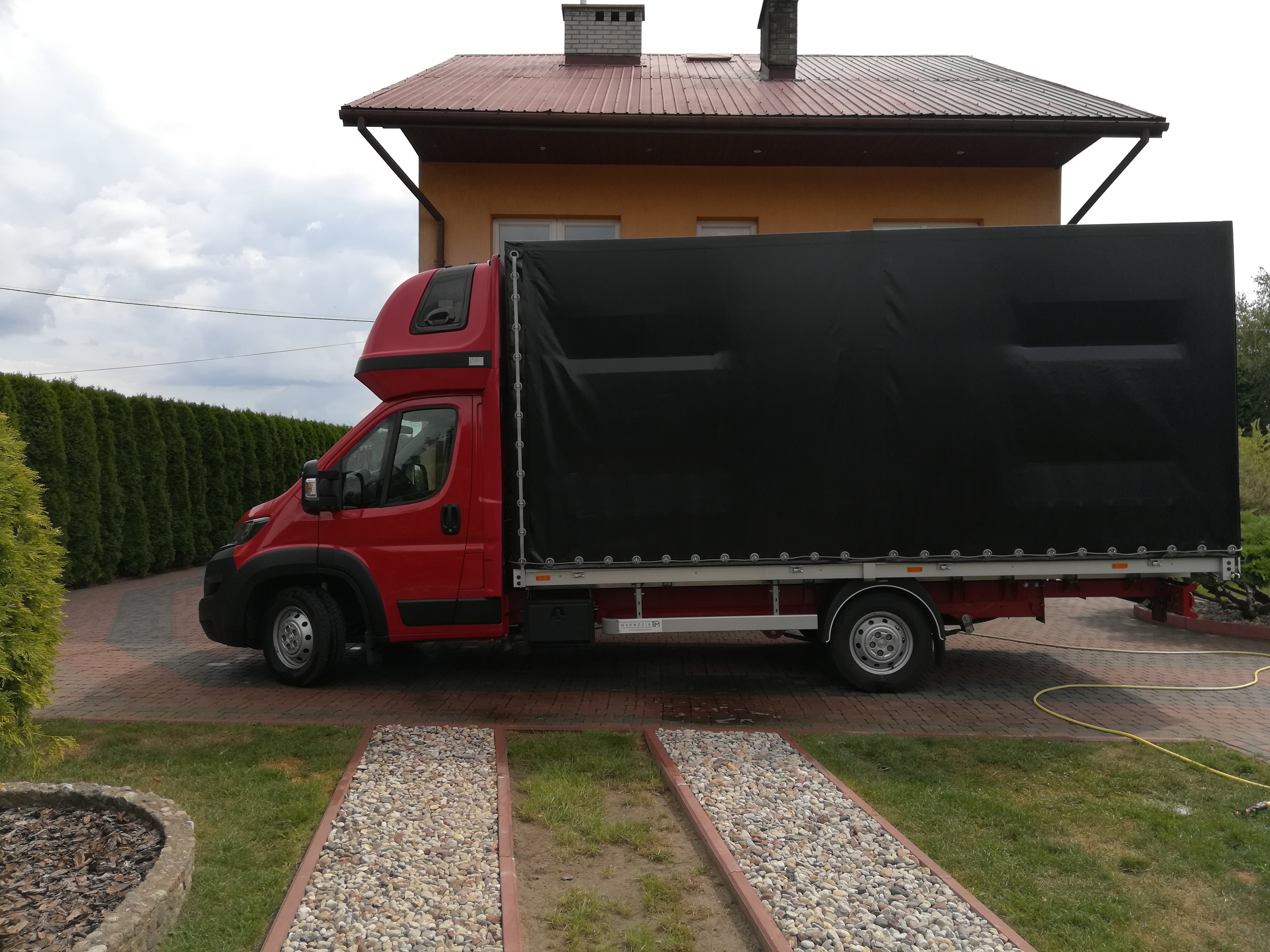 Czerwony bus z czarną plandeką zaparkowany na posesji w Broku, widok z boku, na tle domu z brązowym dachem i zielonego żywopłotu.