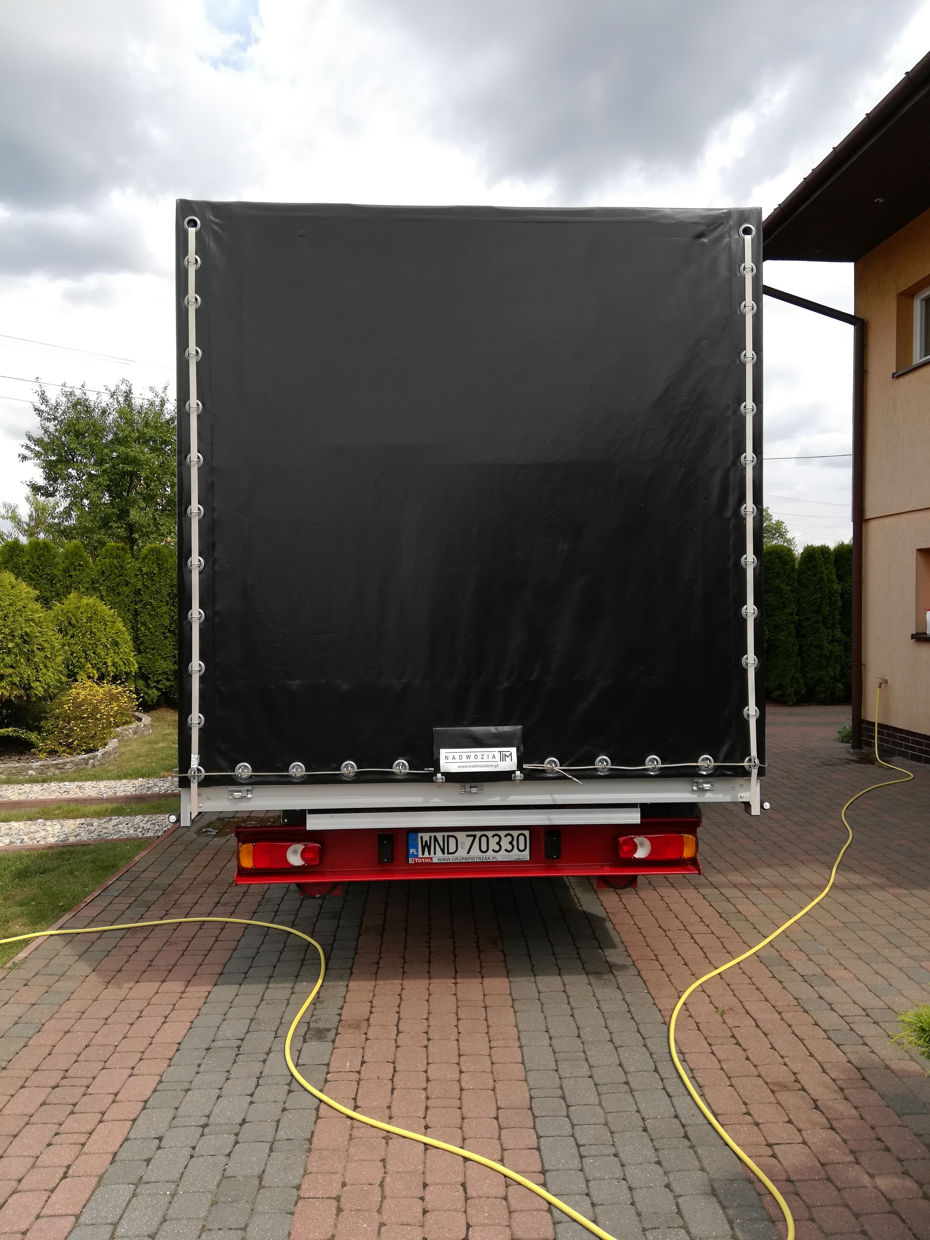Tył samochodu dostawczego z czarną plandeką, widoczny numer rejestracyjny WND 70330 i żółty wąż leżący na kostce brukowej.