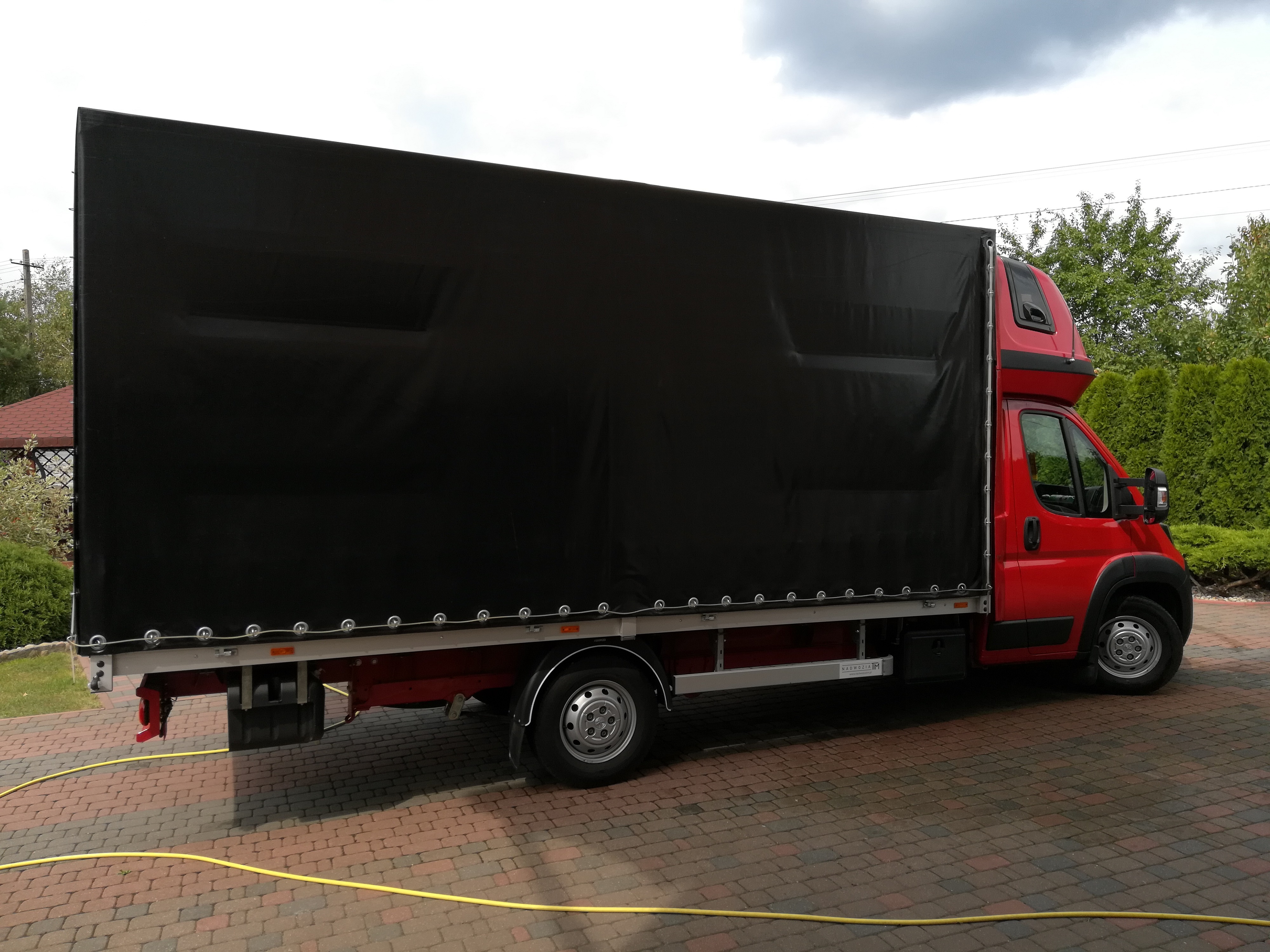 Czerwony samochód dostawczy z czarną plandeką na tle brukowanej nawierzchni i zieleni.