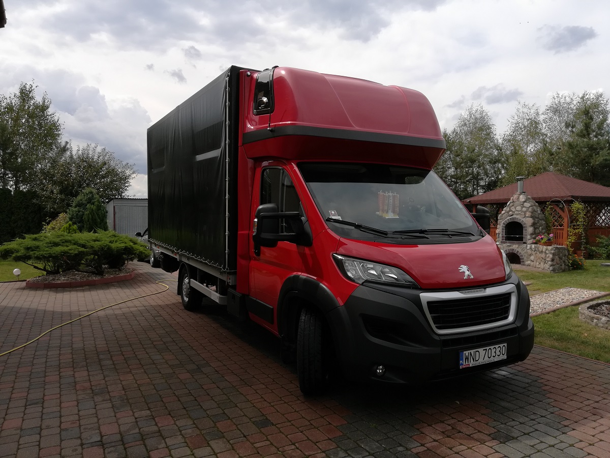 Czerwony bus dostawczy z plandeką na posesji z kostki brukowej, w tle altana z kamiennym grillem.