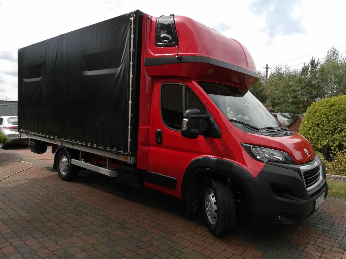 Czerwony samochód dostawczy z czarną plandeką na kostce brukowej, widoczny fragment zaparkowanego samochodu osobowego w tle.