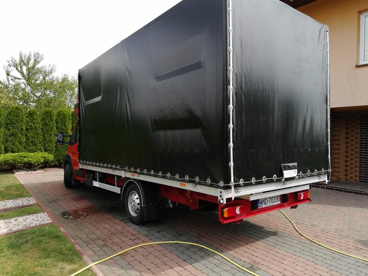 Czerwony bus z czarną plandeką, zaparkowany na kostce brukowej przed domem, widok z tyłu, tablica rejestracyjna WND 70330.