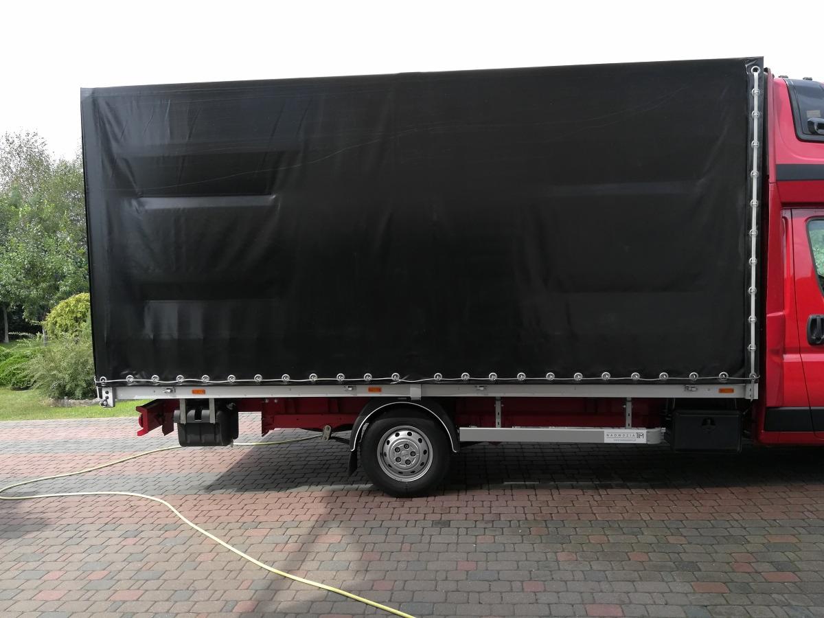 Czerwony bus z czarną plandeką na naczepie, widok z boku na kostce brukowej, z widocznym przewodem.