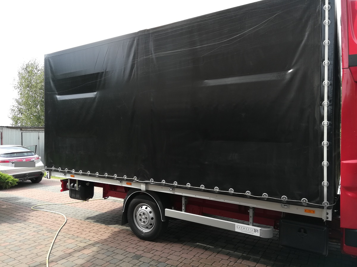 Czerwony bus z czarną plandeką na tle kostki brukowej i zaparkowanego samochodu osobowego.