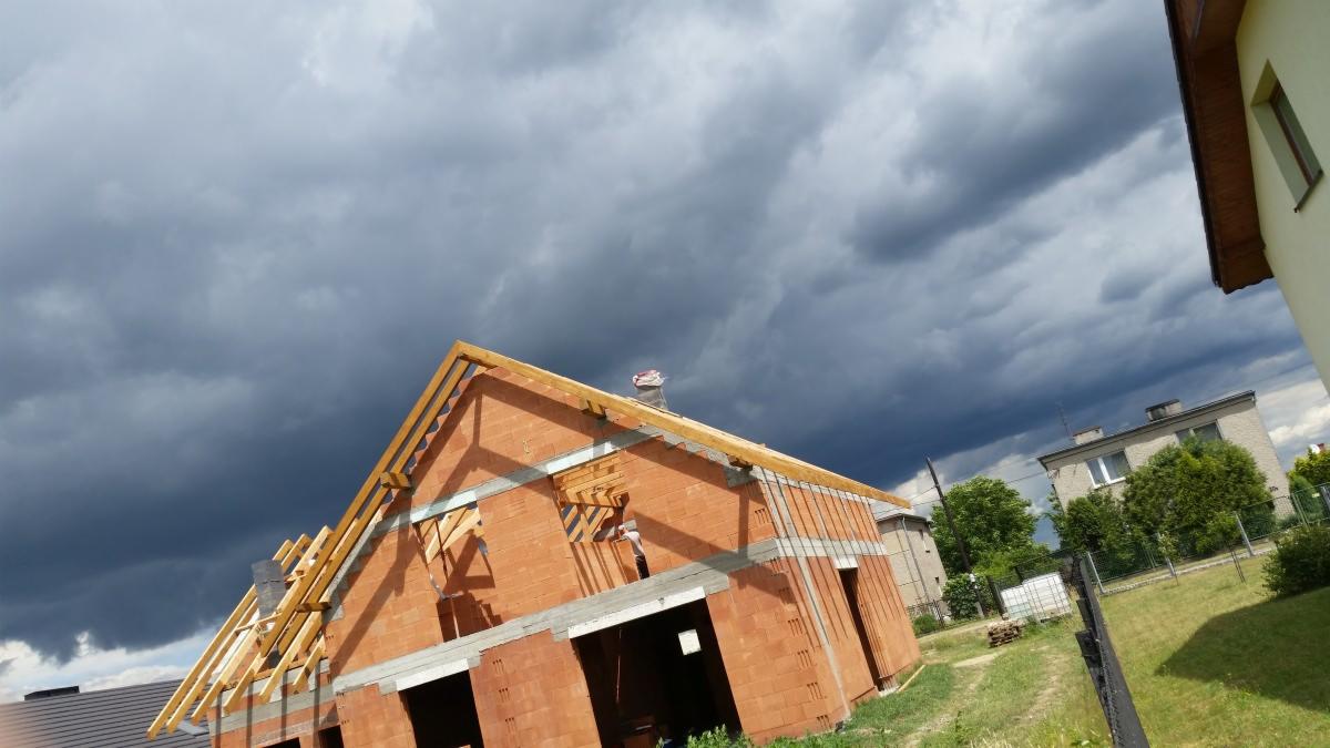 Niedokończony dom z czerwonej cegły pod burzliwym niebem; widoczne drewniane elementy konstrukcji dachu i metalowe wzmocnienia ścian.