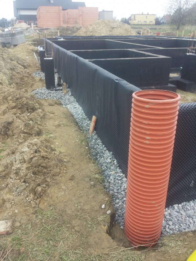 Fundamenty domu jednorodzinnego w trakcie budowy, zabezpieczone folią kubełkową i drenażem, widoczna pomarańczowa rura kanalizacyjna.