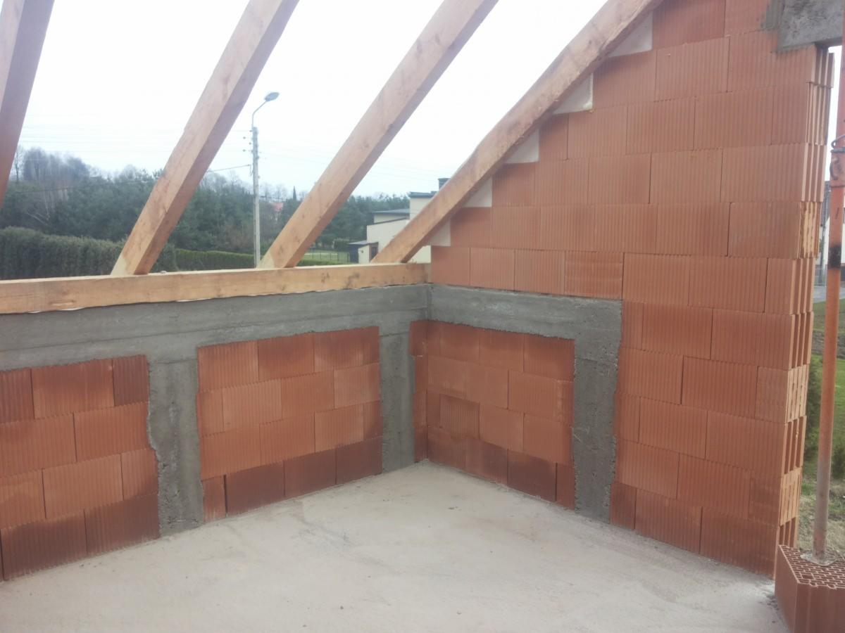 Wnętrze surowego domu murowanego w trakcie budowy, widoczne ściany z czerwonej cegły ceramicznej, betonowe nadproża i drewniana konstrukcja dachu, na zewnątrz zieleń drzew.