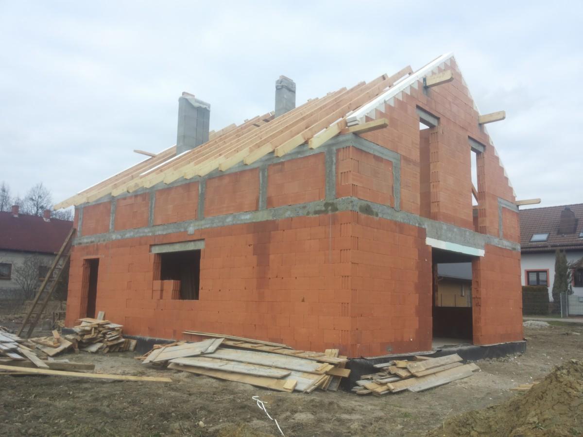 Surowy stan dwukondygnacyjnego domu z czerwonej cegły z widoczną konstrukcją dachu z drewna, kominami i nieotynkowanymi ścianami w trakcie budowy.