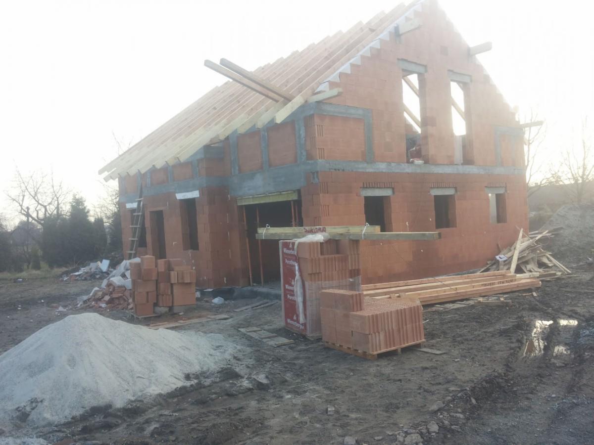 Surowy stan domu jednorodzinnego z czerwonej cegły, widoczne drewniane elementy konstrukcji dachu oraz stosy cegieł i drewna na placu budowy, w tle drzewa bez liści.