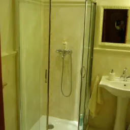 Narożna kabina prysznicowa z chromowanymi profilami i wzorem na płytkach, widoczna umywalka na postumencie z baterią oraz lustro z odbiciem osoby robiącej zdjęcie.