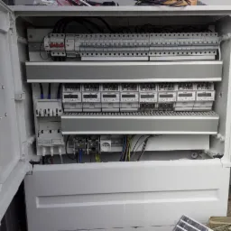 Otwarta, biała skrzynka elektryczna z zamontowanymi licznikami, bezpiecznikami i okablowaniem, widoczne elementy instalacji elektrycznej wewnątrz.