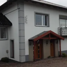 Dwukondygnacyjny dom jednorodzinny z jasną elewacją, szarą podmurówką i ciemnym dachem, widoczny balkon z kutą balustradą i wejście z drewnianym zadaszeniem.