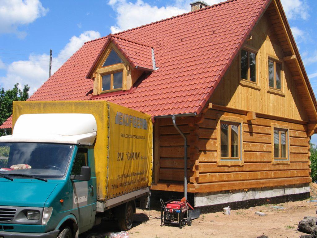 Drewniany dom z czerwoną dachówką, częściowo zasłonięty przez żółty samochód dostawczy z reklamą firmy ociepleniowej. Widoczny generator prądu i fragment ogrodu.