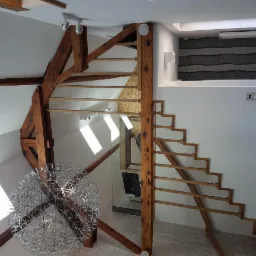 Wnętrze poddasza z drewnianą konstrukcją stropu, schodami bez stopni i nowoczesnym żyrandolem kulistym, oświetlone światłem wpadającym przez okna dachowe.