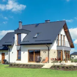 Nowoczesny dom jednorodzinny z ciemną, metalową dachówką, jasną elewacją częściowo wykończoną kamieniem, widoczny taras z meblami ogrodowymi i zadbany trawnik.