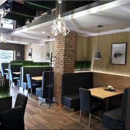 F.H.U. MGM-DESIGN Marcin Guba - Wnętrze restauracji z drewnianymi stołami, czarnymi lożami i krzesłami, ceglanym filarem i dekoracyjnym oświetleniem.