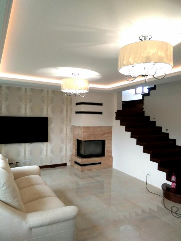 Jasne wnętrze salonu z kominkiem obłożonym marmurem, kremową sofą, błyszczącą podłogą z płytek, dwoma lampami sufitowymi z abażurami i schodami z ciemnego drewna.