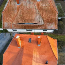 Widok z góry na dwa dachy z czerwonej dachówki. Górny dach jest częściowo pokryty mchem i zabrudzeniami, a dolny dach jest czysty i świeżo wyglądający. Na dolnym dachu widać postać w ciemnym ubraniu.