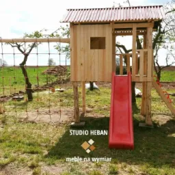 Drewniany plac zabaw z domkiem, zjeżdżalnią i siatką wspinaczkową na tle zielonego trawnika i drzew, z logo firmy meblowej w rogu.