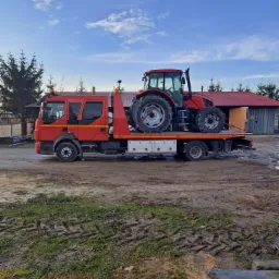Czerwona ciężarówka z platformą transportową przewozi czerwony traktor na tle budynku gospodarczego i drzew iglastych.