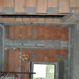 Wnętrze w trakcie budowy z widocznymi przewodami elektrycznymi rozprowadzonymi po ścianach z cegły i betonu, puszki instalacyjne zamontowane, okno w tle.