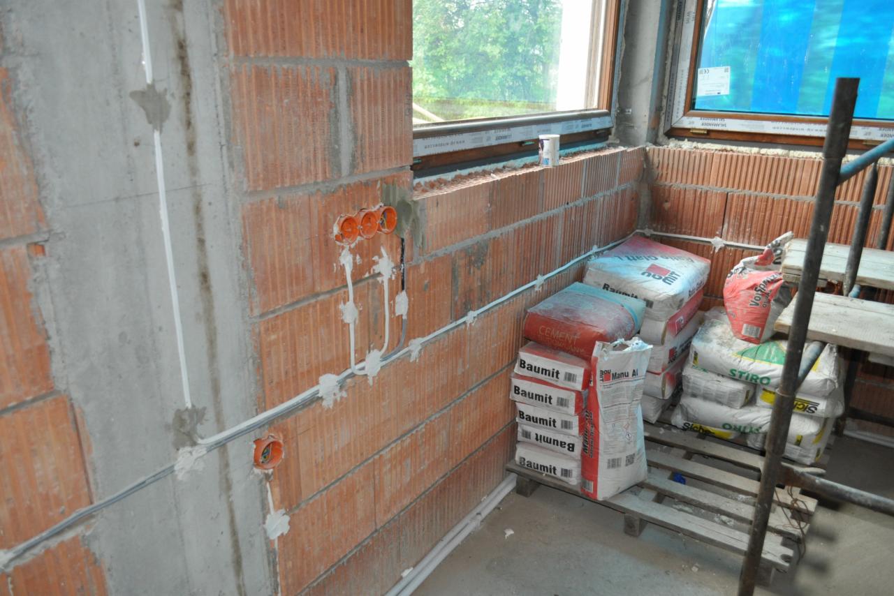 Surowy mur z cegły z widocznymi puszkami instalacyjnymi i przewodami elektrycznymi, częściowo otynkowany, na tle okna i worków z materiałami budowlanymi.