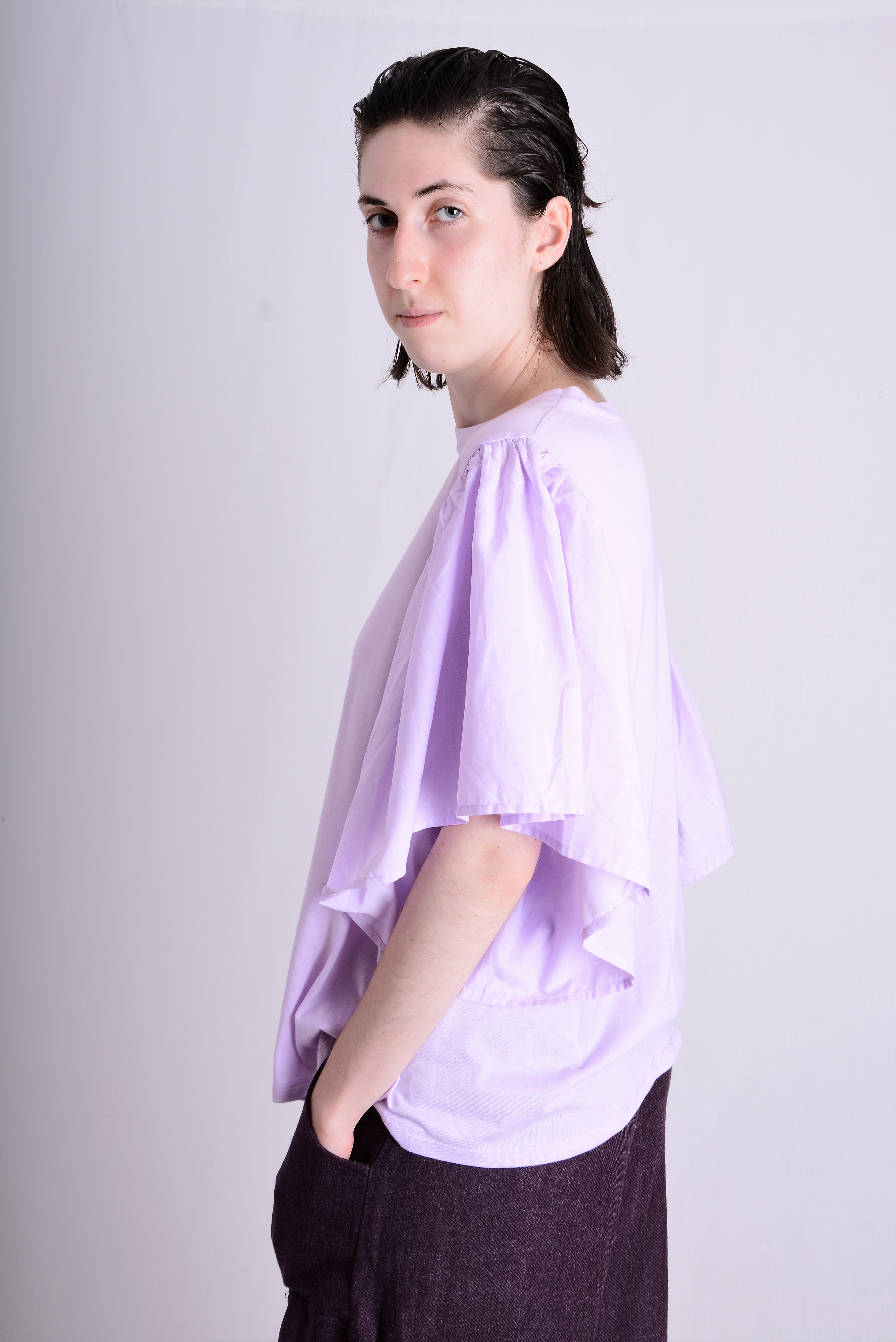 Portret kobiety w liliowej bluzce z rękawami typu motyl, pozującej na jasnym tle, z ręką w kieszeni ciemnych spodni