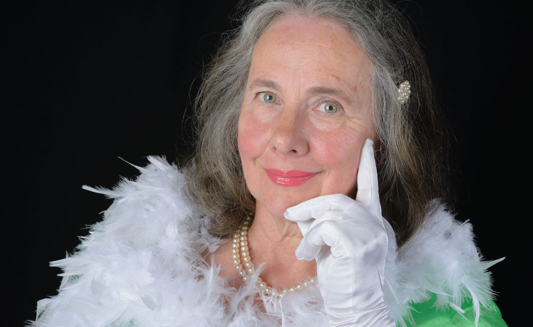 Portret kobiety w białych rękawiczkach i boa z piór, z perłami na szyi i spinką we włosach, pozującej na czarnym tle.