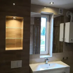 Nowoczesna łazienka z brązowymi płytkami, wbudowaną niszą z szklanymi półkami, podwieszaną szafką z umywalką i dużym lustrem odbijającym okno.