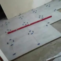 Układanie szarych płytek podłogowych z użyciem poziomicy i niebieskich krzyżyk&oacute;w dystansowych, widoczne niedociągnięcia przy ścianie i fragment obuwia.