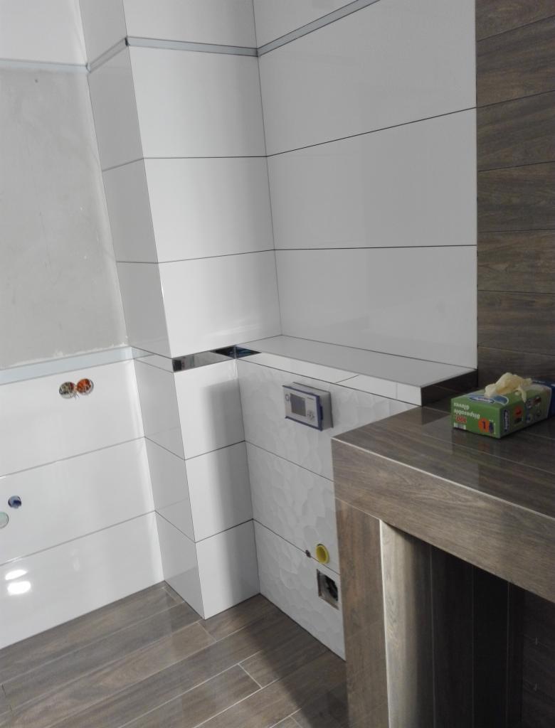 Nowoczesna łazienka po remoncie: białe, poziome płytki z metalowymi listwami, imitacja drewna na ścianie i podłodze, widoczne przyłącza hydrauliczne i elektryczne, pudełko rękawic na blacie.