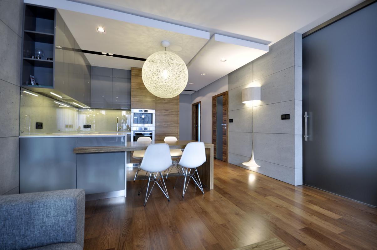 Nowoczesna kuchnia z jadalnią w szarościach i drewnie: szare szafki, drewniany blat, białe krzesła Eames, okrągła lampa wisząca z włókna, drewniana podłoga i przesuwne drzwi.