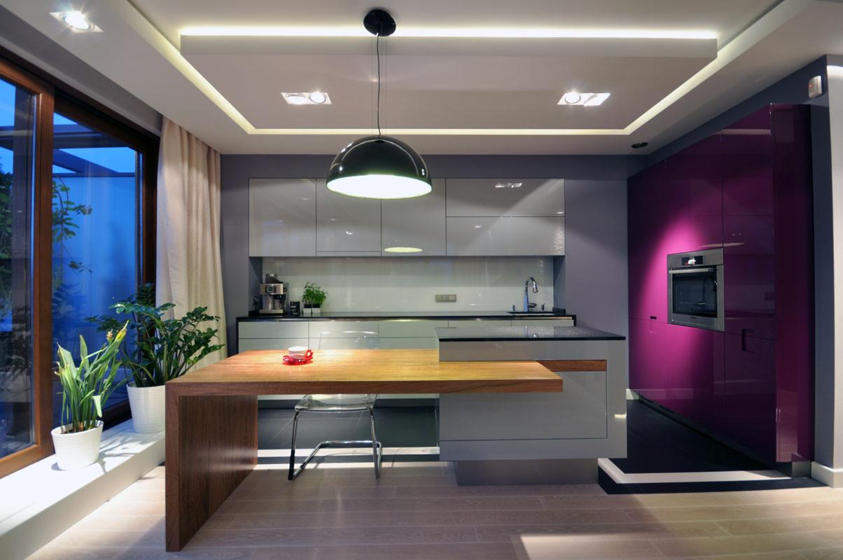 Nowoczesna kuchnia z drewnianym stołem-barem, przezroczystym krzesłem, szarymi szafkami i fioletową ścianą z piekarnikiem w zabudowie, oświetlona wiszącą lampą i ledowym oświetleniem sufitowym.